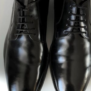 May include: A pair of polished black leather shoes with black laces. The shoes have a classic design and a glossy finish, suitable for formal events. The image shows the shoes from a top-down perspective.