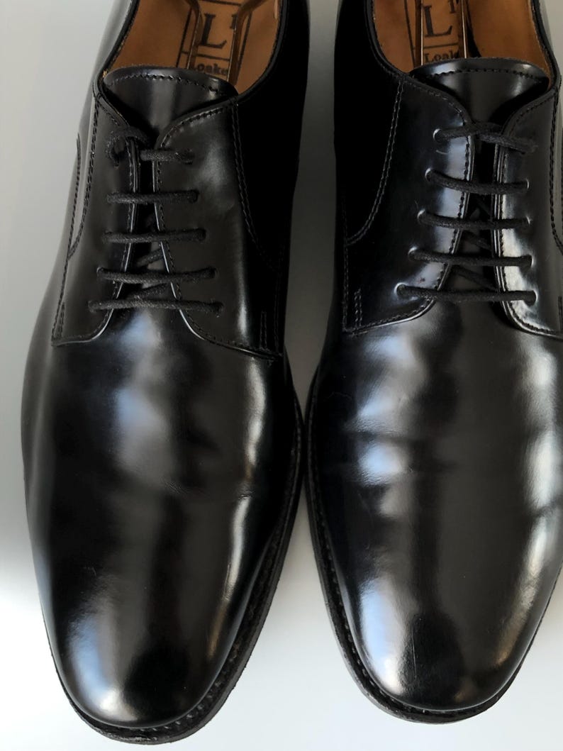 May include: A pair of polished black leather shoes with black laces. The shoes have a classic design and a glossy finish, suitable for formal events. The image shows the shoes from a top-down perspective.