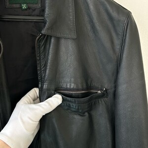 May include: A black leather jacket featuring a classic collar, a full-length front zipper, and a zippered chest pocket. The interior label displays the brand name SPRINGFIELD with size information. The jacket has a smooth texture and long sleeves.