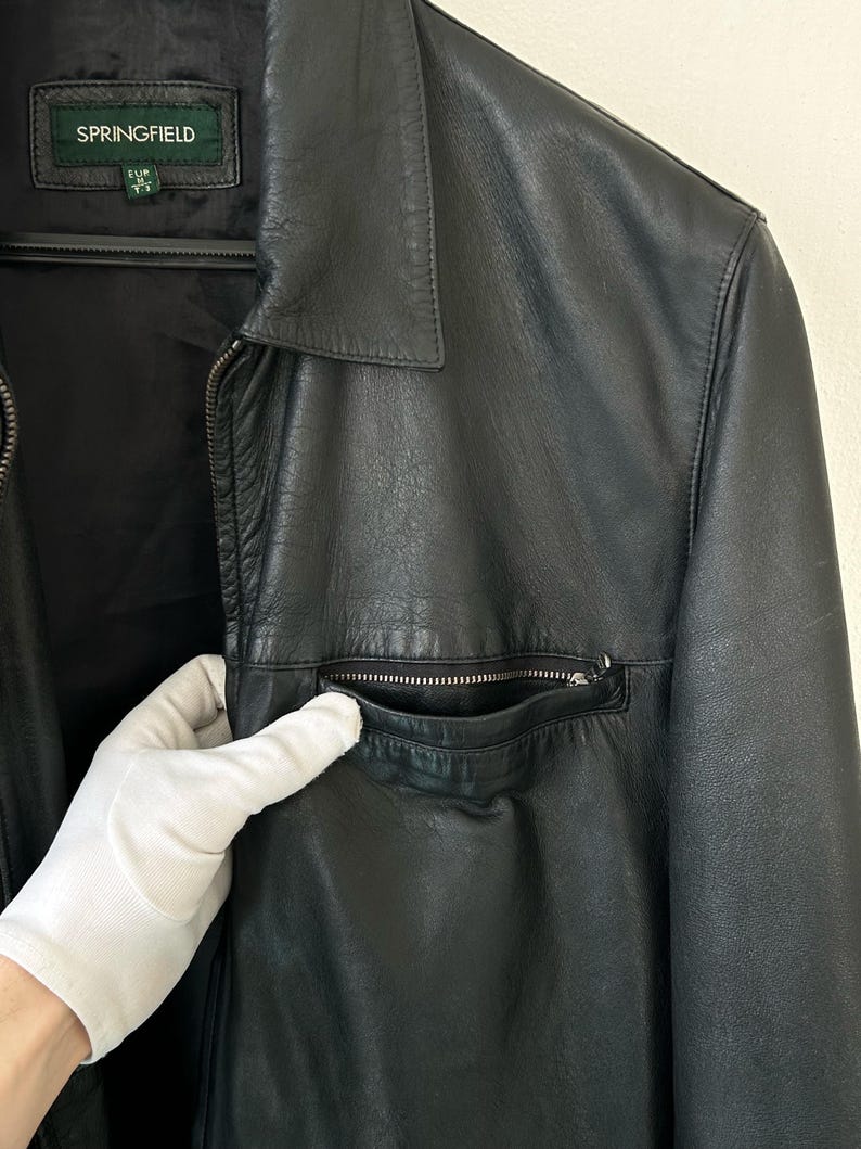 May include: A black leather jacket featuring a classic collar, a full-length front zipper, and a zippered chest pocket. The interior label displays the brand name SPRINGFIELD with size information. The jacket has a smooth texture and long sleeves.