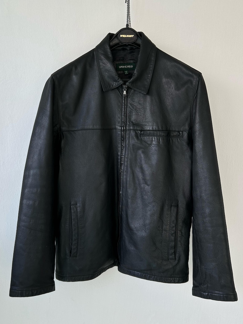 May include: A black jacket with a full-length front zipper, a standard collar, and long sleeves. The jacket features two lower side pockets and one horizontal chest pocket. The interior label displays the brand name Springfield in white text.