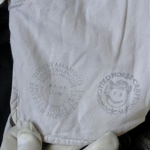 Puede incluir: Primer plano de una tela de color claro con dos logotipos circulares impresos. Un logotipo presenta el texto "YOHJI YAMAMOTO" y el otro presenta una cara de dibujos animados y el texto "SPOTTED HORSE CRAFT WASH".