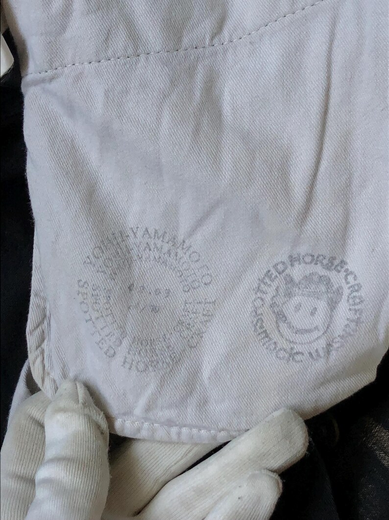 Puede incluir: Primer plano de una tela de color claro con dos logotipos circulares impresos. Un logotipo presenta el texto "YOHJI YAMAMOTO" y el otro presenta una cara de dibujos animados y el texto "SPOTTED HORSE CRAFT WASH".