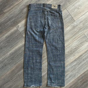 May include: A pair of blue denim jeans featuring a textured, cross-hatched pattern throughout the fabric. The jeans include two back pockets, a waistband with belt loops, and a rectangular Emporio Armani brand patch on the back right side.