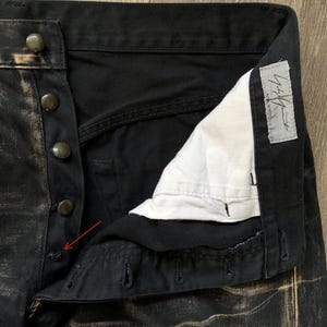 Puede incluir: Primer plano de unos pantalones vaqueros negros con bragueta de botones. El tejido vaquero tiene un aspecto desgastado y descolorido con detalles en color beige. Los pantalones tienen botones oscuros y un forro interior blanco. Se ve una etiqueta con texto japon&eacute;s.
