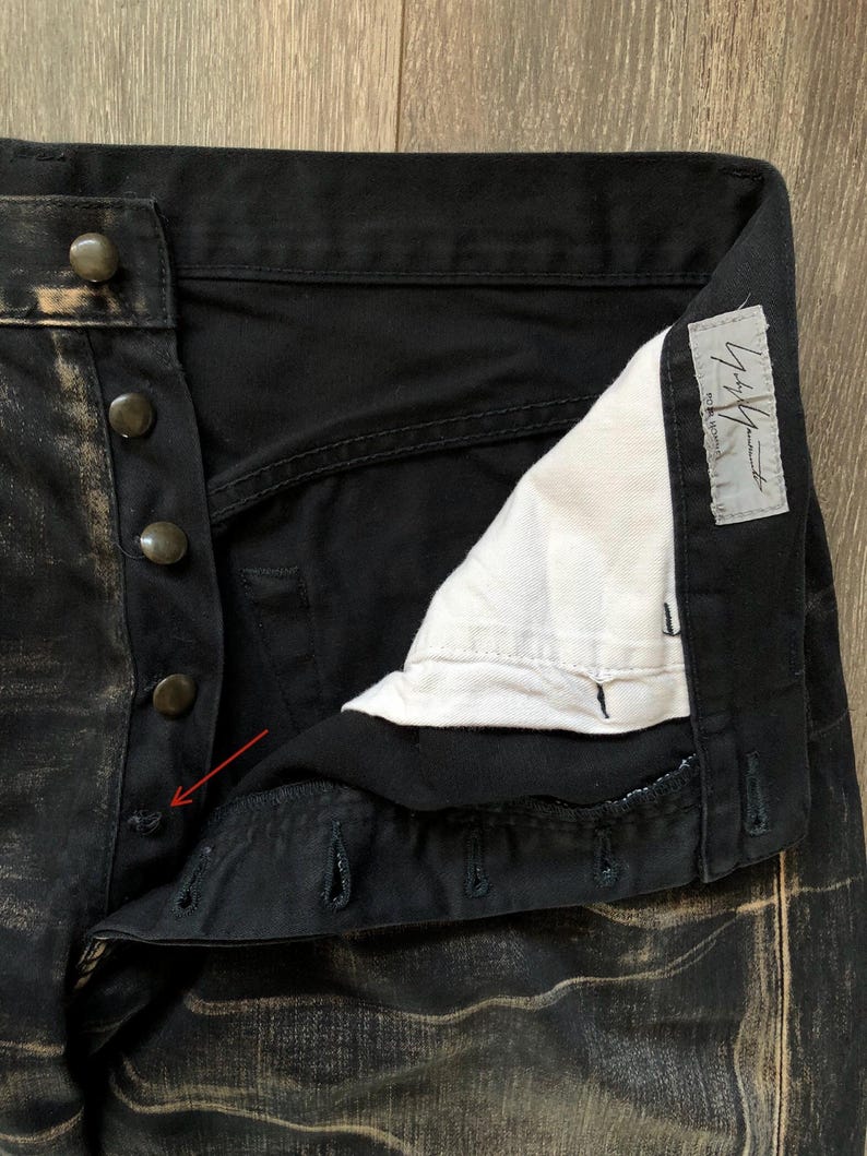 Puede incluir: Primer plano de unos pantalones vaqueros negros con bragueta de botones. El tejido vaquero tiene un aspecto desgastado y descolorido con detalles en color beige. Los pantalones tienen botones oscuros y un forro interior blanco. Se ve una etiqueta con texto japon&eacute;s.