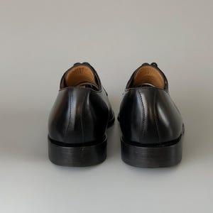 May include: A pair of black leather dress shoes, seen from behind. The shoes have a polished finish, visible stitching, and a stacked heel. The interior of the shoe is a light brown colour.
