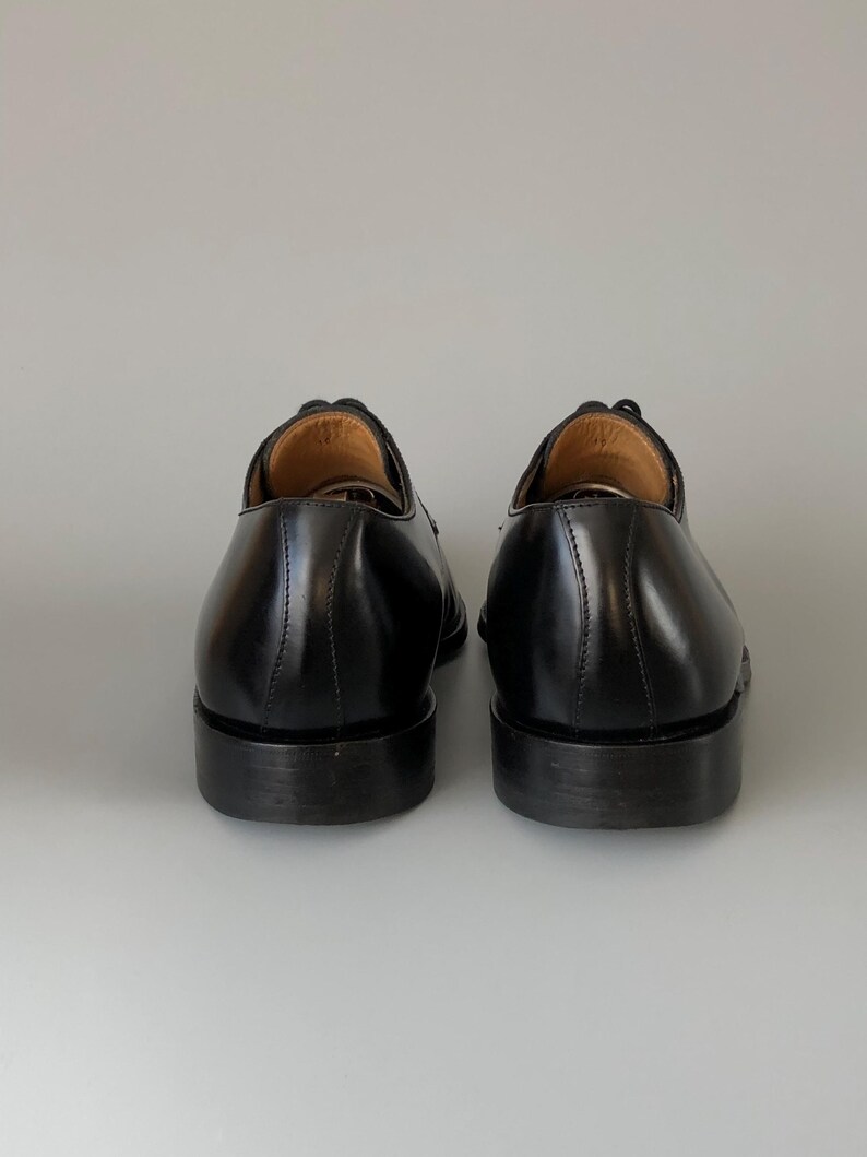 May include: A pair of black leather dress shoes, seen from behind. The shoes have a polished finish, visible stitching, and a stacked heel. The interior of the shoe is a light brown colour.