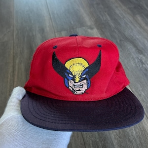 Puede incluir: Una gorra de béisbol roja con visera azul oscuro que presenta un gráfico bordado del personaje de Marvel Wolverine en el panel frontal. El diseño del personaje incluye una máscara amarilla y negra con detalles en azul y un pequeño símbolo TM.