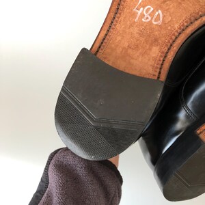 May include: Close-up of a black leather shoe, showing the sole and heel. The sole is light brown with stitching and the number "480" in white. The heel is black rubber with a textured pattern. The shoe appears worn.
