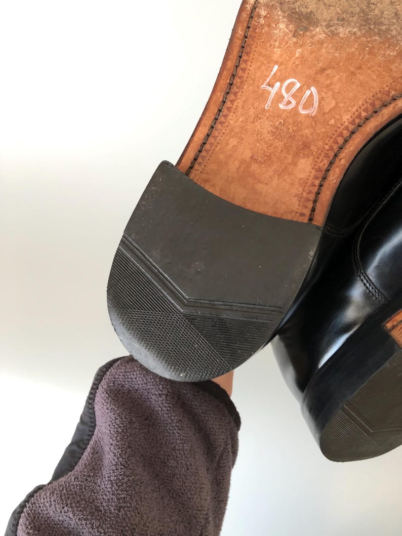 May include: Close-up of a black leather shoe, showing the sole and heel. The sole is light brown with stitching and the number "480" in white. The heel is black rubber with a textured pattern. The shoe appears worn.
