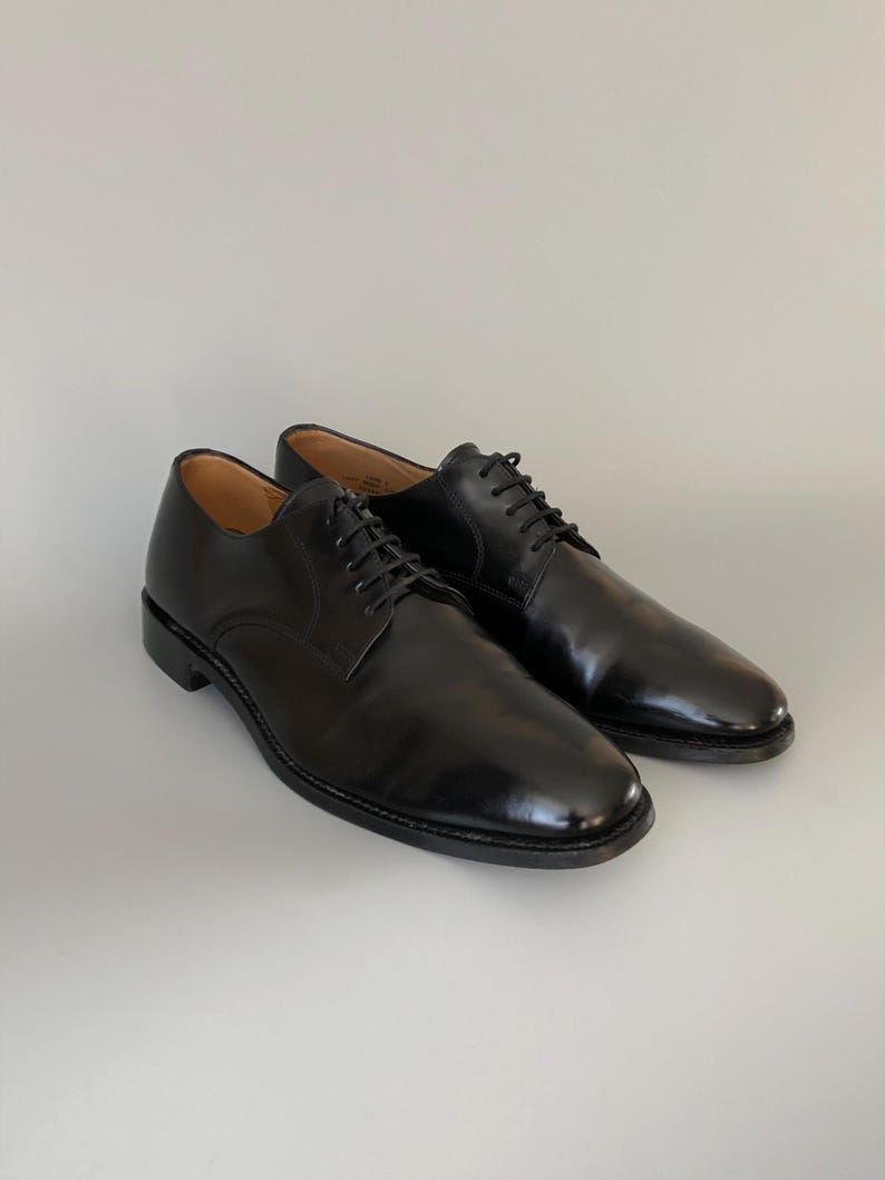 May include: A pair of black leather dress shoes with laces. The shoes have a glossy finish and a classic design, suitable for formal occasions. The shoes are displayed on a white surface.