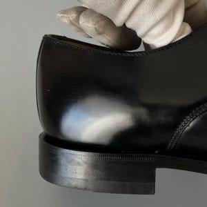 May include: Close-up of a black leather shoe, highlighting the polished surface and stitching. The heel is visible, with a textured sole. The image focuses on the craftsmanship and design of the footwear.