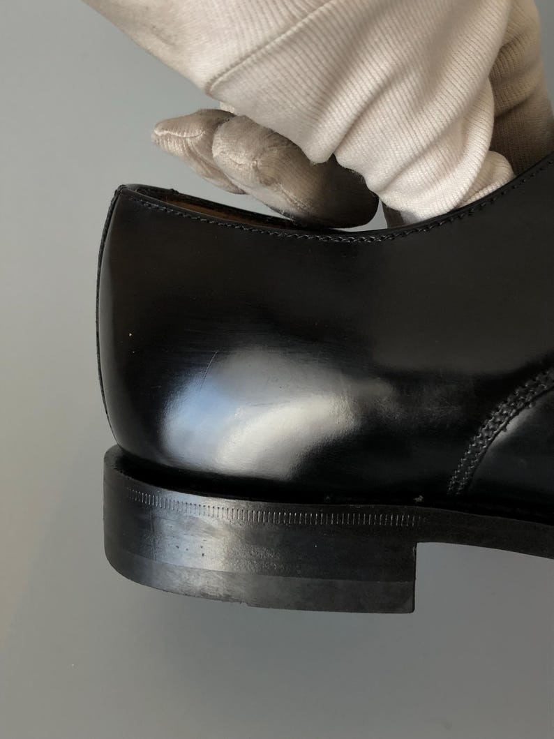 May include: Close-up of a black leather shoe, highlighting the polished surface and stitching. The heel is visible, with a textured sole. The image focuses on the craftsmanship and design of the footwear.