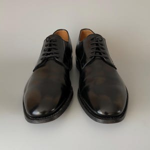 May include: Two black leather dress shoes with a high-shine finish and a traditional lace-up closure. The shoes have a rounded toe and a dark sole. The image shows the shoes against a plain background, suitable for formal wear.