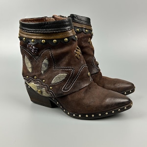 May include: A pair of brown ankle boots featuring a pointed toe, a stacked heel, and decorative brass studs along the sole and collar. The boots are crafted from dark brown suede with lighter textured leaf-shaped overlays and a black leather trim.