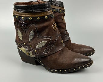 Puede incluir: Un par de botines marrones con punta afilada, tacón de bloque y tachuelas decorativas de latón a lo largo de la suela y el cuello. Las botas están hechas de ante marrón oscuro con superposiciones texturizadas en forma de hoja y ribete de cuero negro.