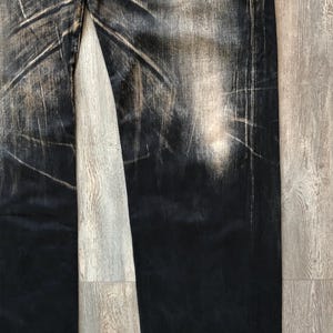 Puede incluir: Primer plano de vaqueros negros desgastados con zonas descoloridas y detalles texturizados. Los pantalones vaqueros tienen un corte recto y muestran signos de desgaste, con acentos marrones claros. El aspecto general es vintage y robusto.