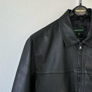 May include: A black leather jacket featuring a classic collar, a full-length front zipper with a silver-toned pull, and a horizontal seam across the chest. The interior label displays the brand name Springfield with size M clearly visible.