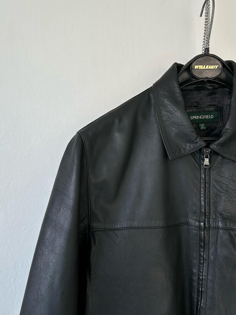 May include: A black leather jacket featuring a classic collar, a full-length front zipper with a silver-toned pull, and a horizontal seam across the chest. The interior label displays the brand name Springfield with size M clearly visible.