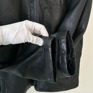 May include: A black leather jacket featuring a smooth texture, long sleeves with finished cuffs, and a central vertical zipper closure. The garment displays visible stitching details along the seams and a structured collar at the top neckline.