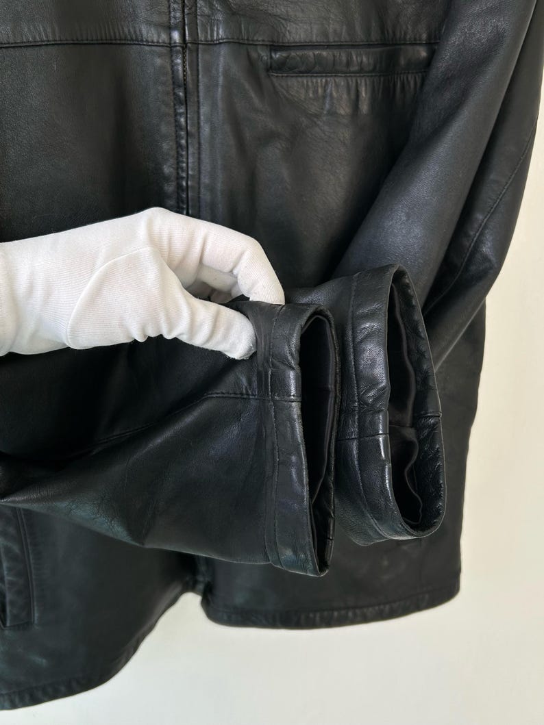 May include: A black leather jacket featuring a smooth texture, long sleeves with finished cuffs, and a central vertical zipper closure. The garment displays visible stitching details along the seams and a structured collar at the top neckline.