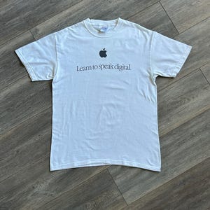 May include: A white short-sleeved crew neck t-shirt featuring a black Apple logo positioned centrally on the chest above the text "Learn to speak digital." The garment is made of cotton and has a standard fit with a ribbed neckline and straight hem.