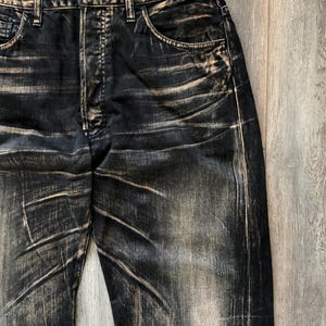 Puede incluir: Primer plano de unos vaqueros de mezclilla negros con un dise&ntilde;o desgastado y descolorido. Los vaqueros tienen un dise&ntilde;o cl&aacute;sico de cinco bolsillos, cierre de bot&oacute;n y trabillas para el cintur&oacute;n. Las zonas descoloridas son de color beige.