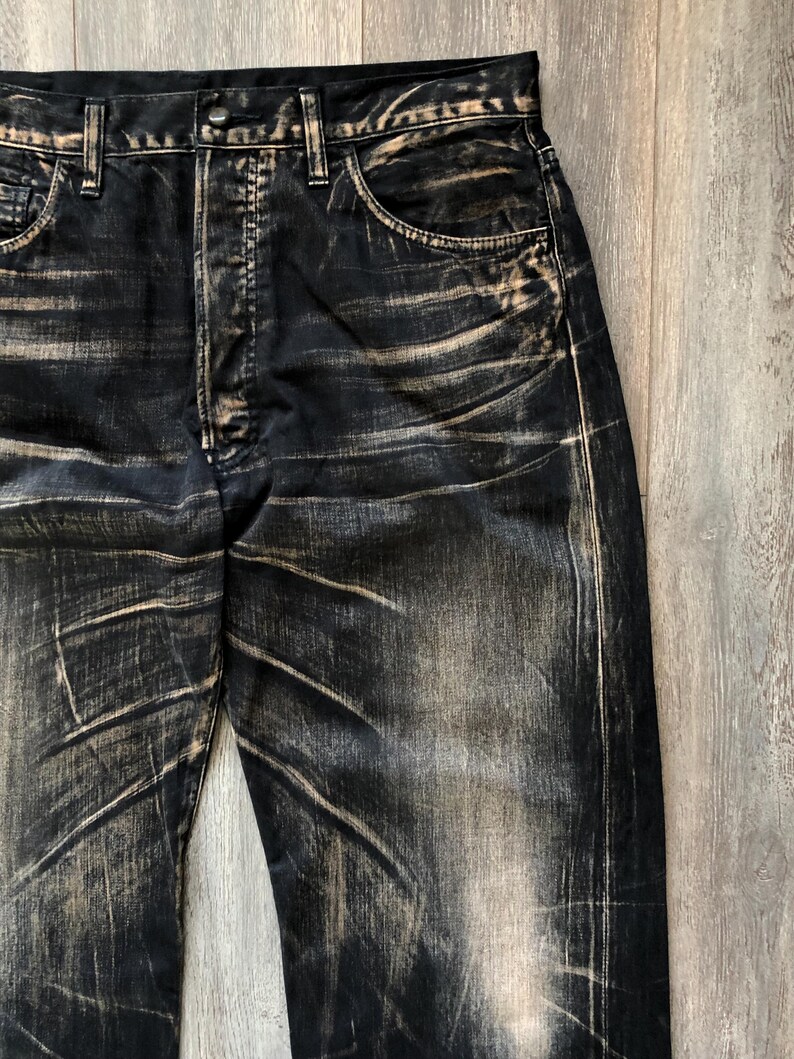 Puede incluir: Primer plano de unos vaqueros de mezclilla negros con un dise&ntilde;o desgastado y descolorido. Los vaqueros tienen un dise&ntilde;o cl&aacute;sico de cinco bolsillos, cierre de bot&oacute;n y trabillas para el cintur&oacute;n. Las zonas descoloridas son de color beige.