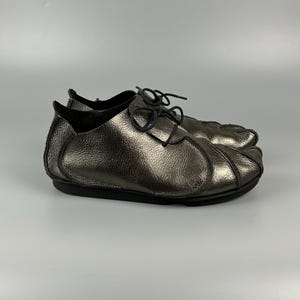 May include: A pair of metallic bronze leather shoes featuring a textured finish, a rounded toe design with stitched paneling, and thin black laces. The shoes have a low-cut silhouette and a flat black sole, providing a sleek and uniform appearance.