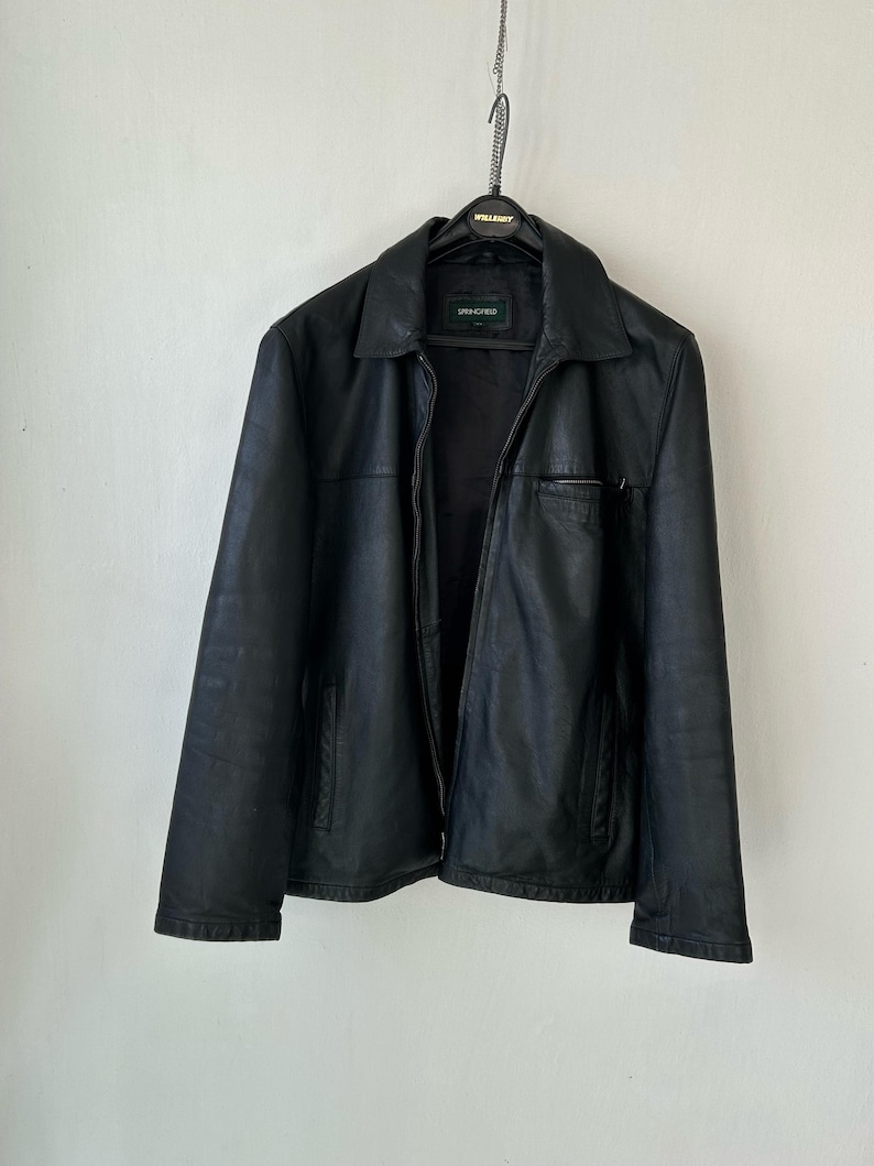 May include: A black leather jacket with a classic collar, a full-length front zipper, and two side pockets. The jacket features a horizontal seam across the chest and a small zippered pocket on the left side. A label reading Springfield is visible.