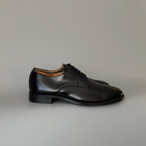 May include: A pair of black leather dress shoes with a polished finish. The shoes feature a classic lace-up design and a low heel. The image displays the shoes from a side perspective, showcasing their elegant shape and shiny appearance.