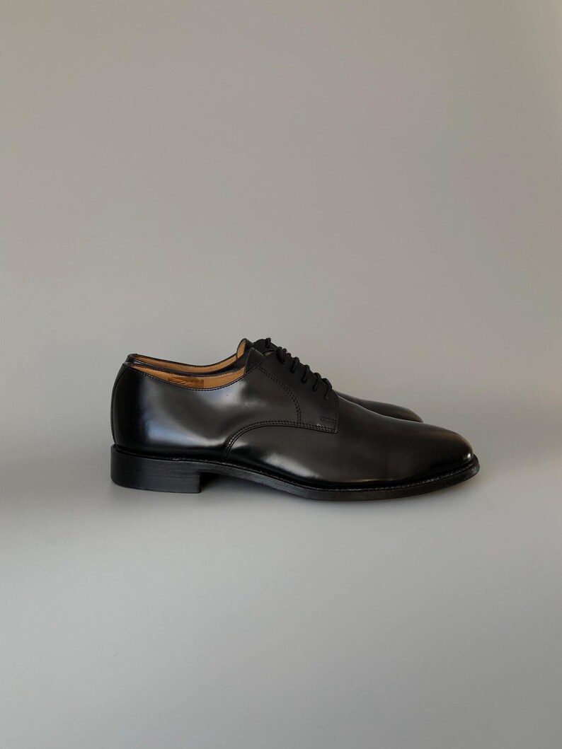May include: A pair of black leather dress shoes with a polished finish. The shoes feature a classic lace-up design and a low heel. The image displays the shoes from a side perspective, showcasing their elegant shape and shiny appearance.