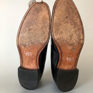 May include: A pair of black leather dress shoes, soles facing up, with the number "480" written in white on the sole. The soles are a light brown colour with visible wear and stitching around the edges.