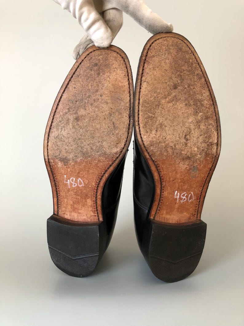 May include: A pair of black leather dress shoes, soles facing up, with the number "480" written in white on the sole. The soles are a light brown colour with visible wear and stitching around the edges.