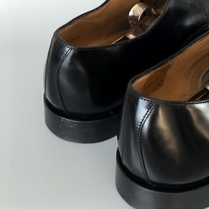 May include: Close-up of two black leather dress shoes. The shoes have a polished finish, visible stitching, and a stacked heel. Shoe trees are inserted inside the shoes. The image highlights the craftsmanship and design of the footwear.