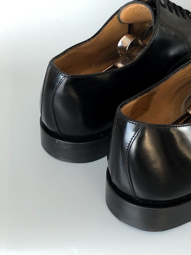 May include: Close-up of two black leather dress shoes. The shoes have a polished finish, visible stitching, and a stacked heel. Shoe trees are inserted inside the shoes. The image highlights the craftsmanship and design of the footwear.