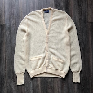Può includere: Un cardigan a maniche lunghe color crema con scollo a V, chiusura frontale con cinque bottoni e due tasche anteriori. Il capo presenta polsini a coste sui polsi e un orlo a coste, realizzato in un tessuto a maglia con una consistenza ruvida.