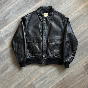 May include: A black leather bomber jacket featuring a classic collar, long sleeves with ribbed cuffs, and a ribbed hem. The jacket includes two large front flap pockets, a central button closure, and a Replay brand label on the inner neck.