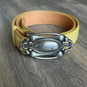 Può includere: Una cintura gialla con un motivo testurizzato simile alla pelle di serpente e una grande fibbia in metallo color argento di forma ovale. La fibbia presenta un design ornato e simmetrico con bordi arrotondati e una finitura centrale opaca, mentre l'interno della cintura è foderato in pelle color cuoio chiaro.
