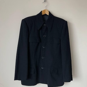 Puede incluir: Blazer negro con cuello, cierre de botones y dos bolsillos delanteros. La chaqueta está colgada en una percha de madera sobre un fondo neutro.