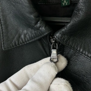 May include: A black leather jacket featuring a pointed collar and a metal zipper pull engraved with the text LAMPO. The interior label displays the brand name SPRINGFIELD and the size label indicates EUR M T-3. The jacket has a textured finish.