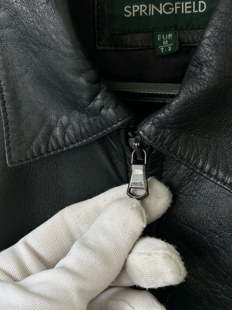 May include: A black leather jacket featuring a pointed collar and a metal zipper pull engraved with the text LAMPO. The interior label displays the brand name SPRINGFIELD and the size label indicates EUR M T-3. The jacket has a textured finish.