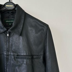 May include: A black leather jacket featuring a classic collar, a full-length front zipper with a metallic pull, and a horizontal chest pocket. The interior label displays the brand name Springfield with size M indicated below the text.