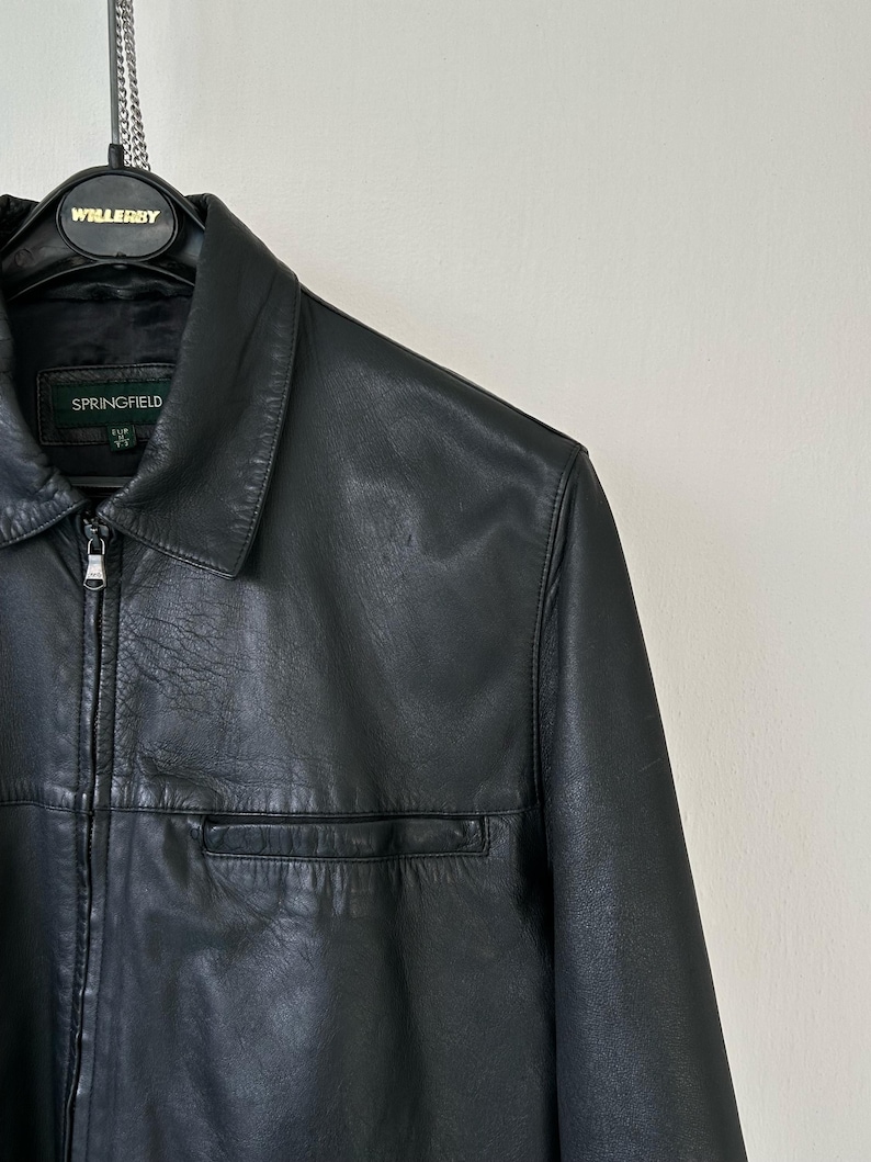 May include: A black leather jacket featuring a classic collar, a full-length front zipper with a metallic pull, and a horizontal chest pocket. The interior label displays the brand name Springfield with size M indicated below the text.