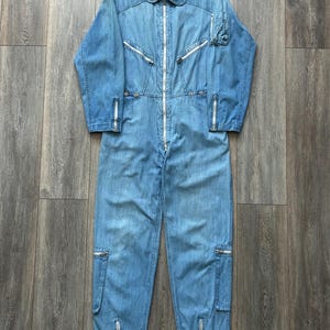 Op de afbeelding: Een lichtblauwe spijkeroverall met een volledige ritssluiting aan de voorzijde, een puntkraag en lange mouwen met ritsmanchetten. Het kledingstuk bevat twee borstzakken met rits, een klein zakje op de mouw en ritsen bij de broekspijpen.