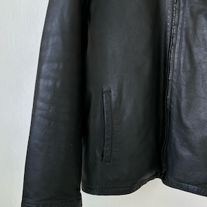 May include: A black leather jacket featuring a smooth texture, a full-length front zipper closure, and two vertical slit pockets on the front. The jacket has long sleeves and a clean, structured design with visible stitching along the seams.