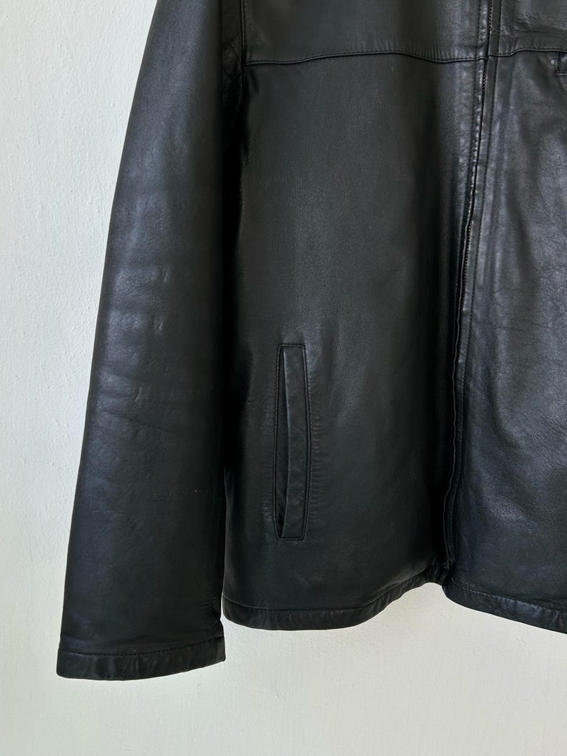 May include: A black leather jacket featuring a smooth texture, a full-length front zipper closure, and two vertical slit pockets on the front. The jacket has long sleeves and a clean, structured design with visible stitching along the seams.