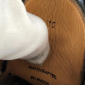 May include: Close-up of the inside of a black leather shoe. The insole is tan with the number "10" printed in black. The words "HANDCRAFTED IN INDIA" are also printed on the insole.