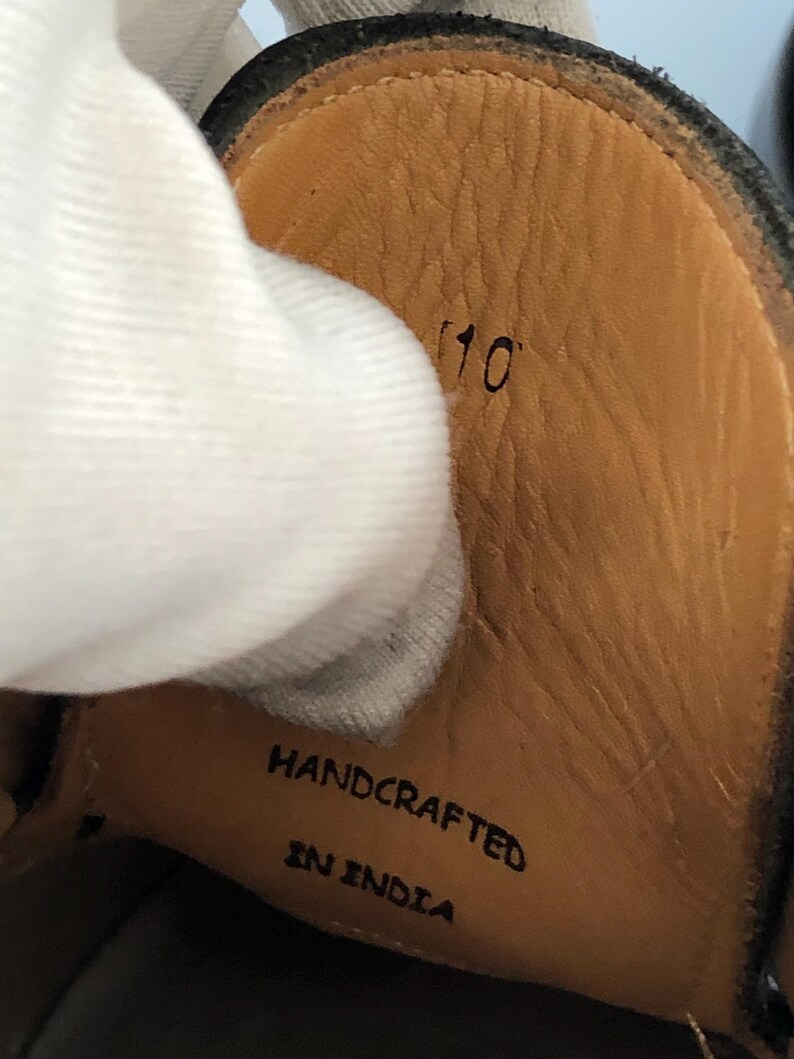 May include: Close-up of the inside of a black leather shoe. The insole is tan with the number "10" printed in black. The words "HANDCRAFTED IN INDIA" are also printed on the insole.